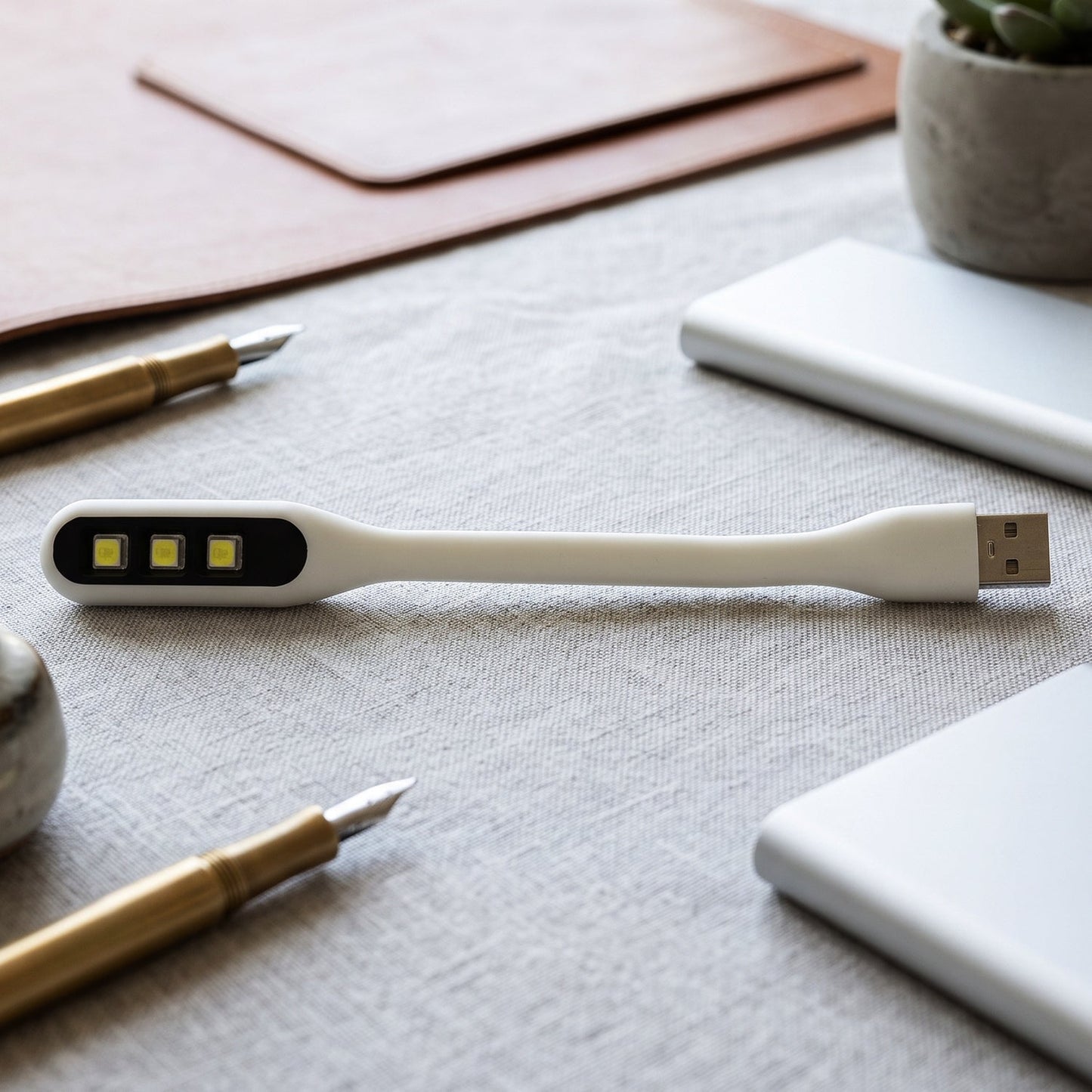 A white flexible USB LED light with three yellow LED bulbs lies on a light gray textured surface surrounded by office items.