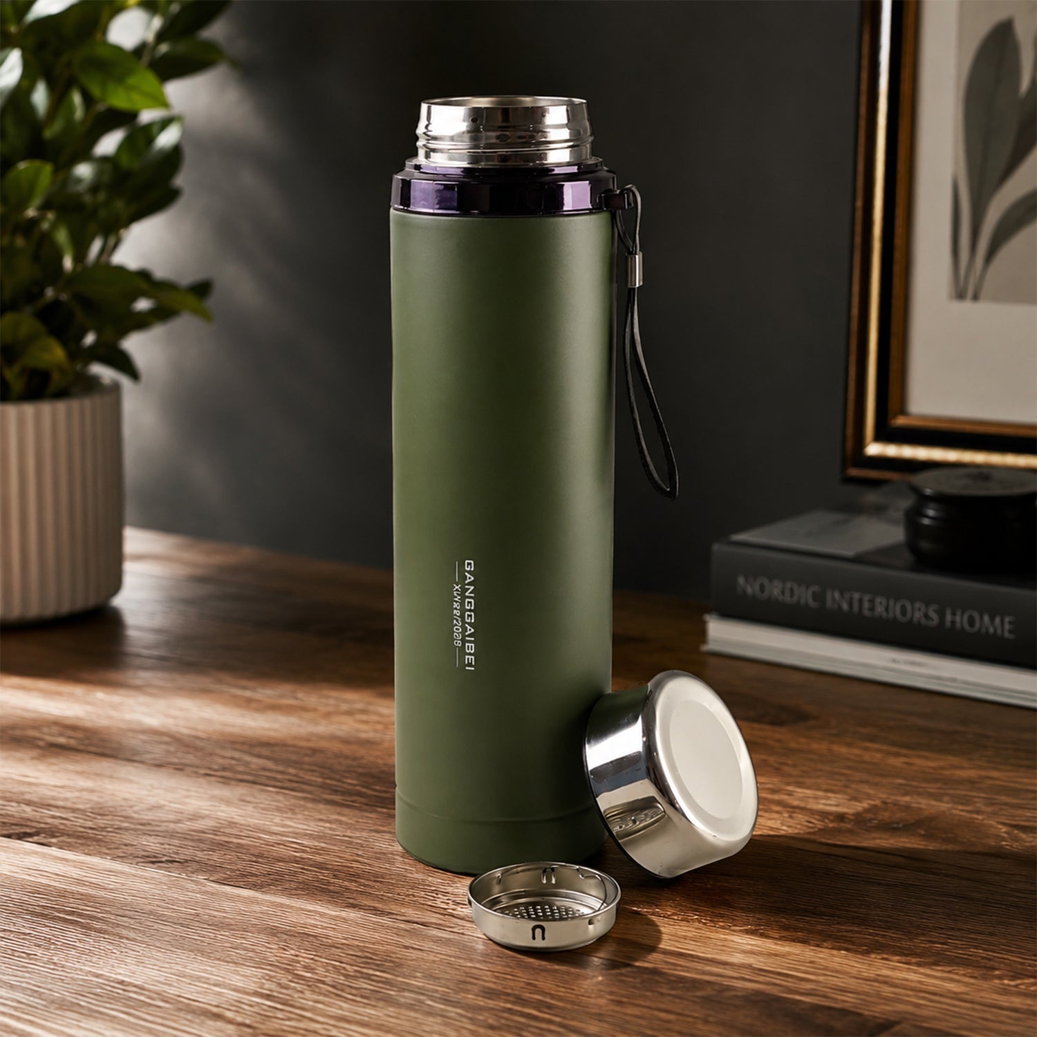 A matte green water bottle with its silver cup lid and infuser detached, on a wooden desk with a plant.