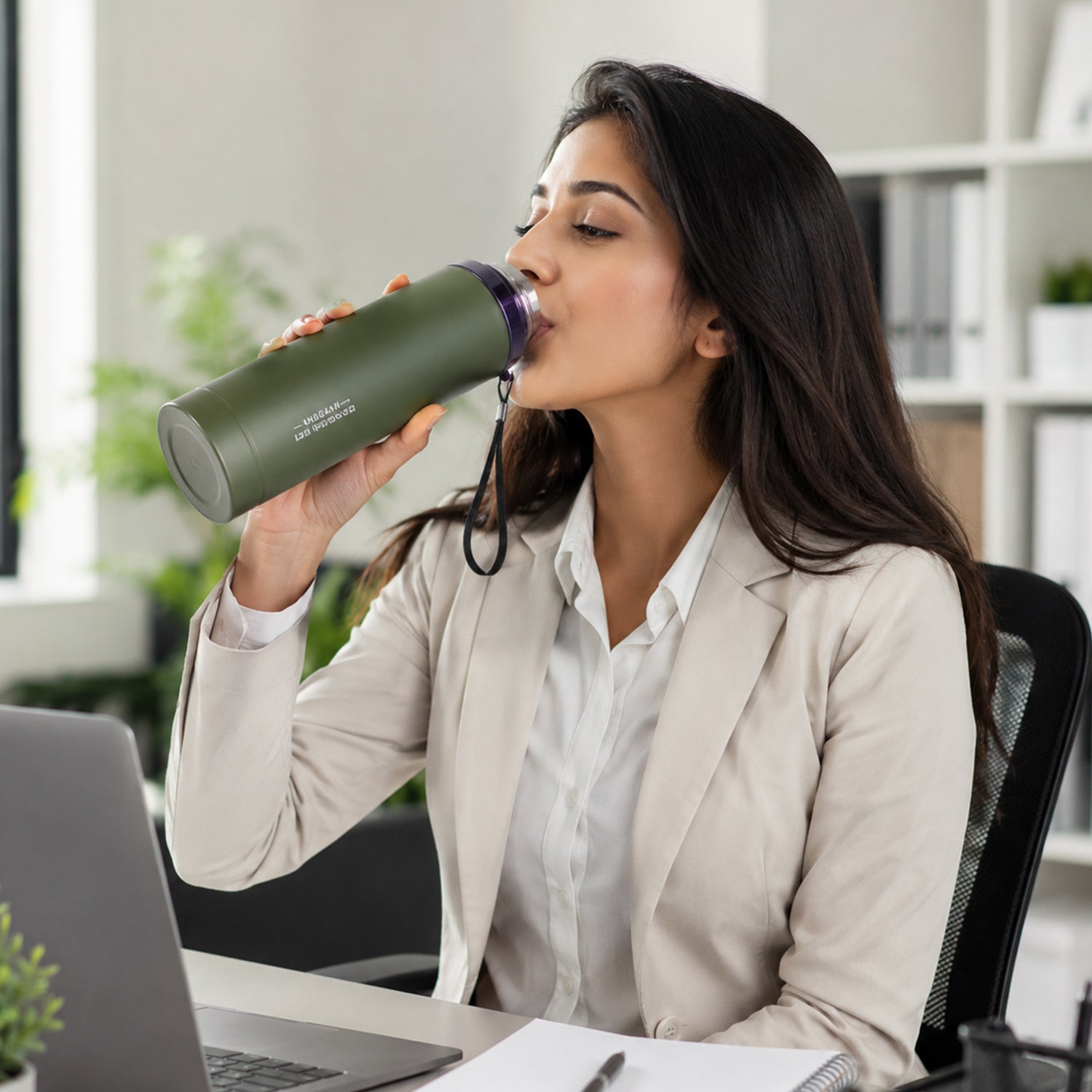 A woman in a beige blazer drinks from a matte green water bottle while sitting at a desk with a laptop.