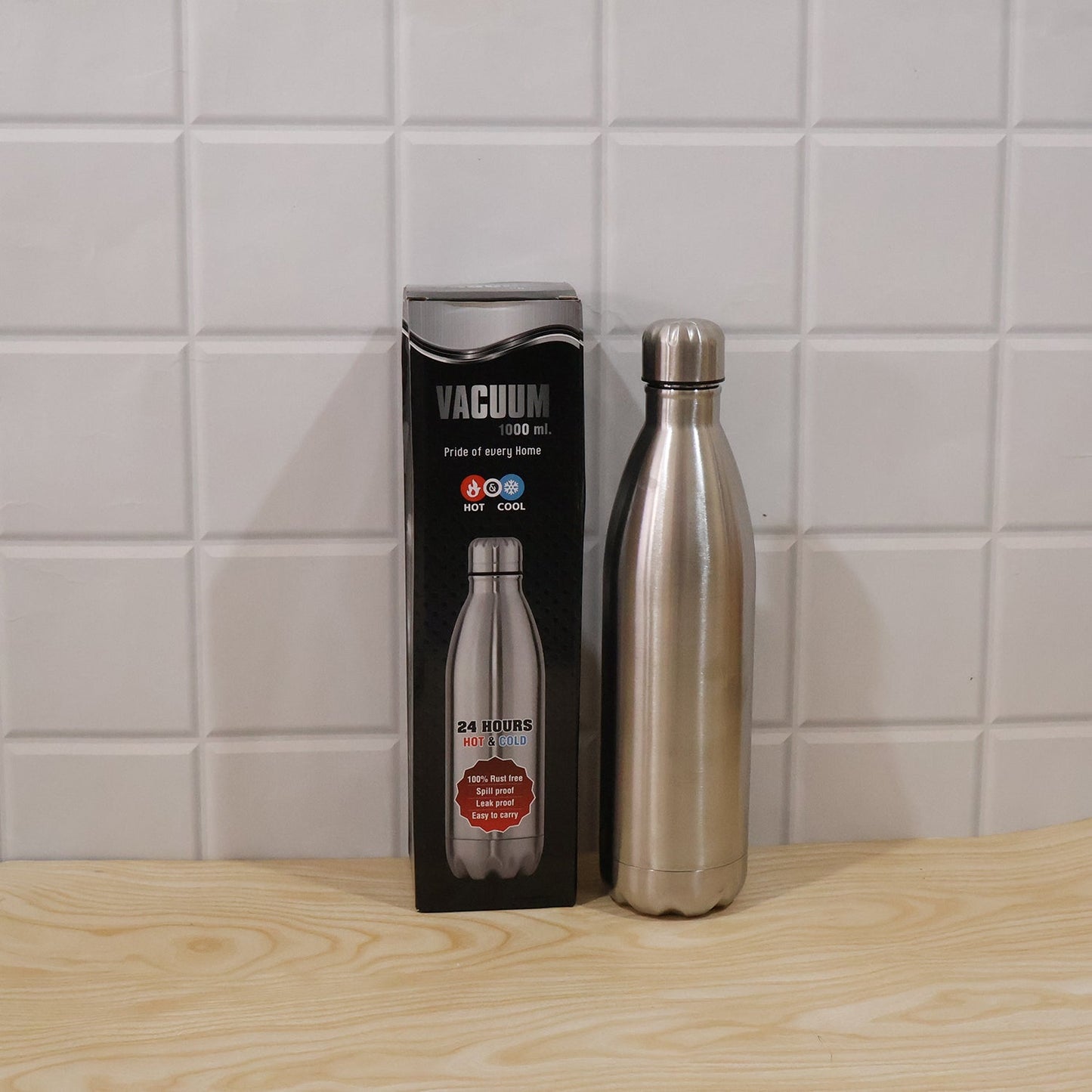 A silver stainless steel water bottle stands next to its black packaging box, both on a wooden surface against a white tiled