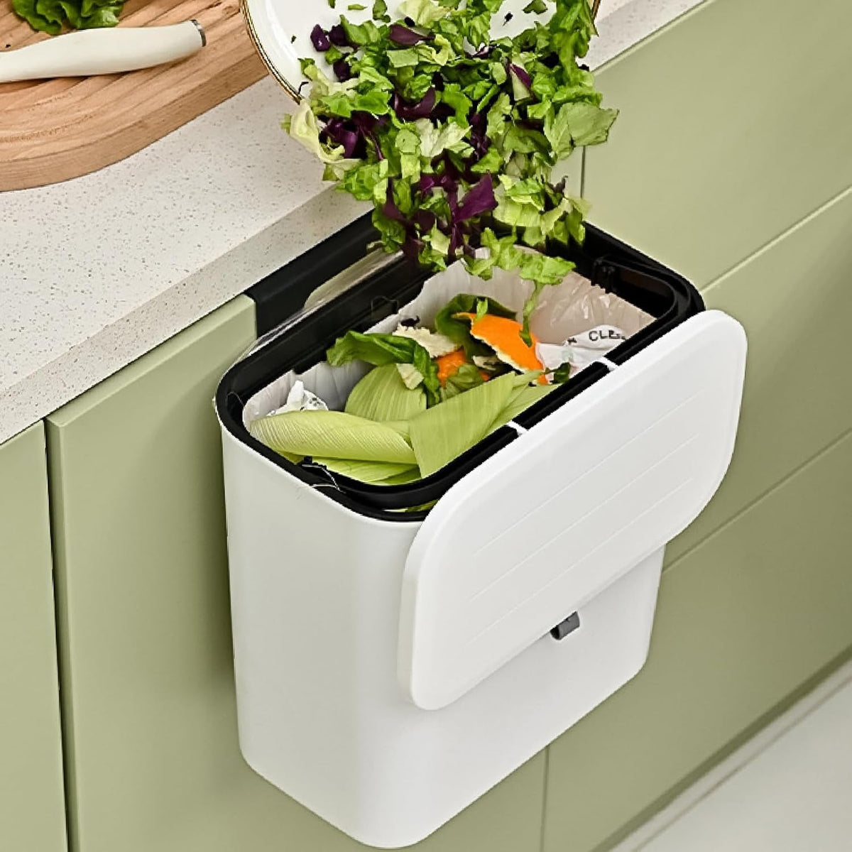 Chopped green vegetables are scraped from a plate into a white wall-mounted compost bin with an open lid.