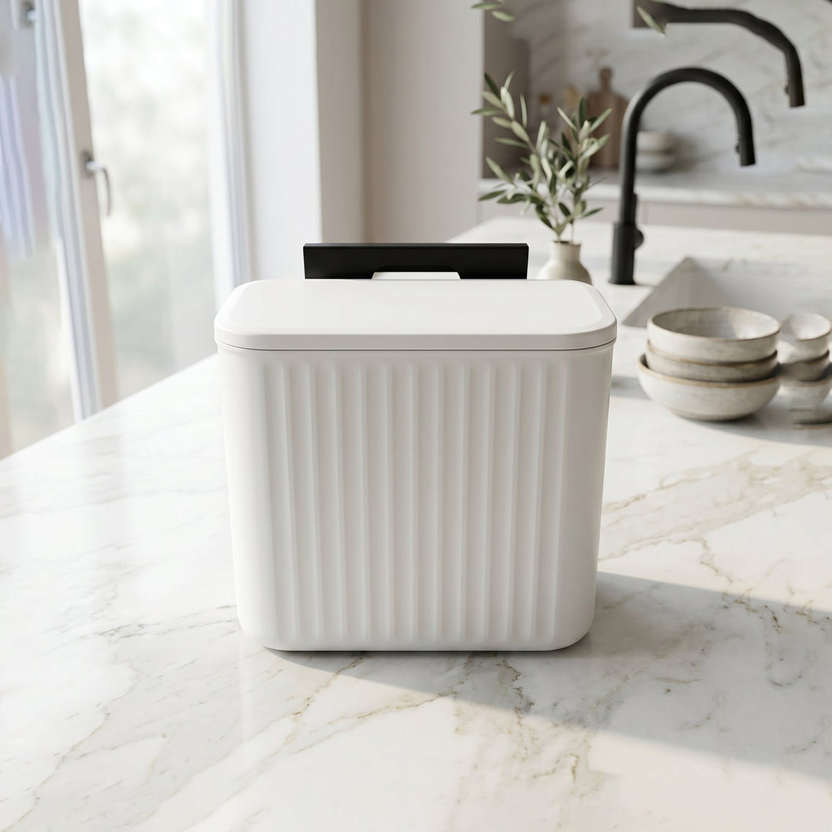 A white ribbed compost bin with a closed lid and black mounting bracket sits on a white marble kitchen counter.