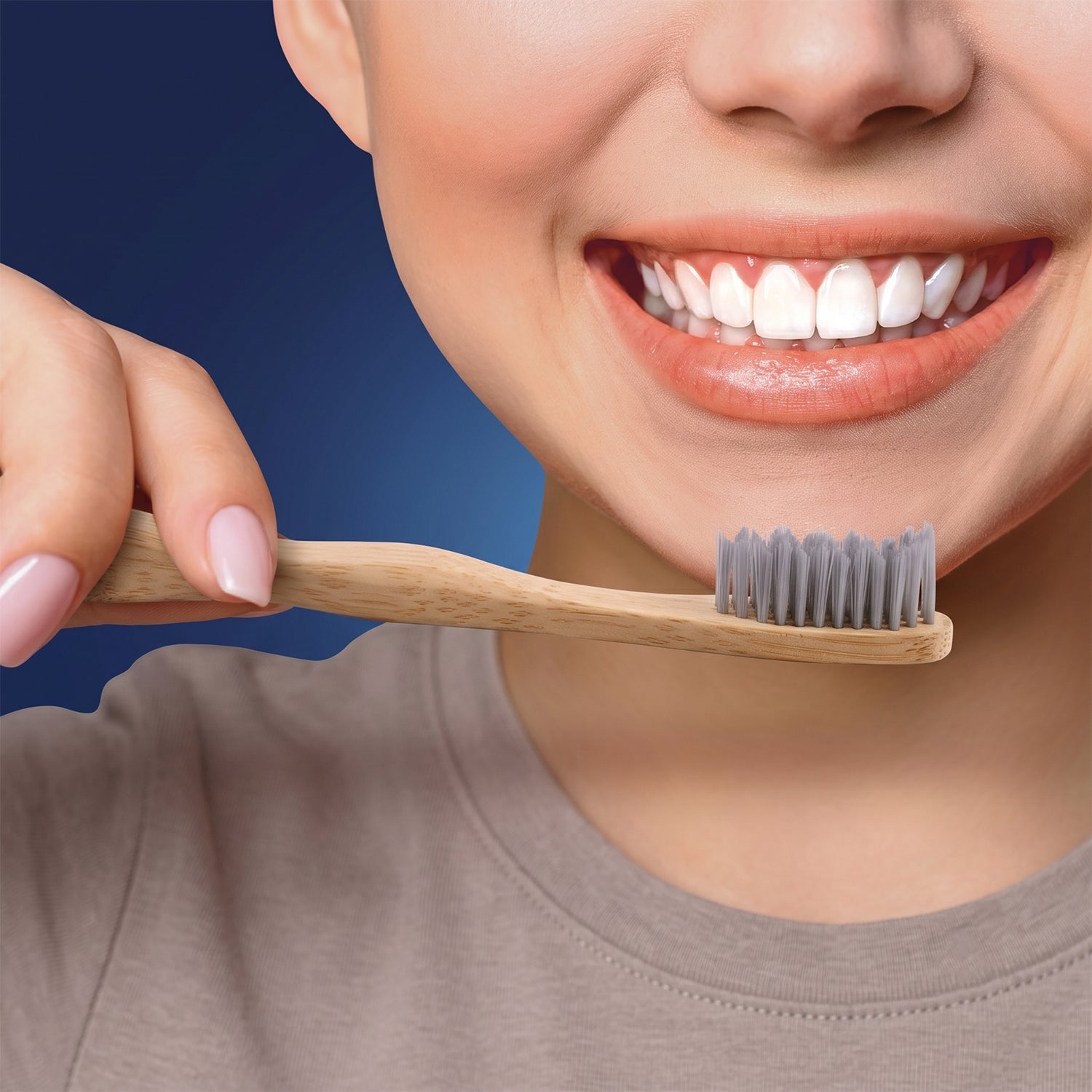 A person with a bright smile holds a bamboo toothbrush with grey bristles near their mouth, against a dark blue background.