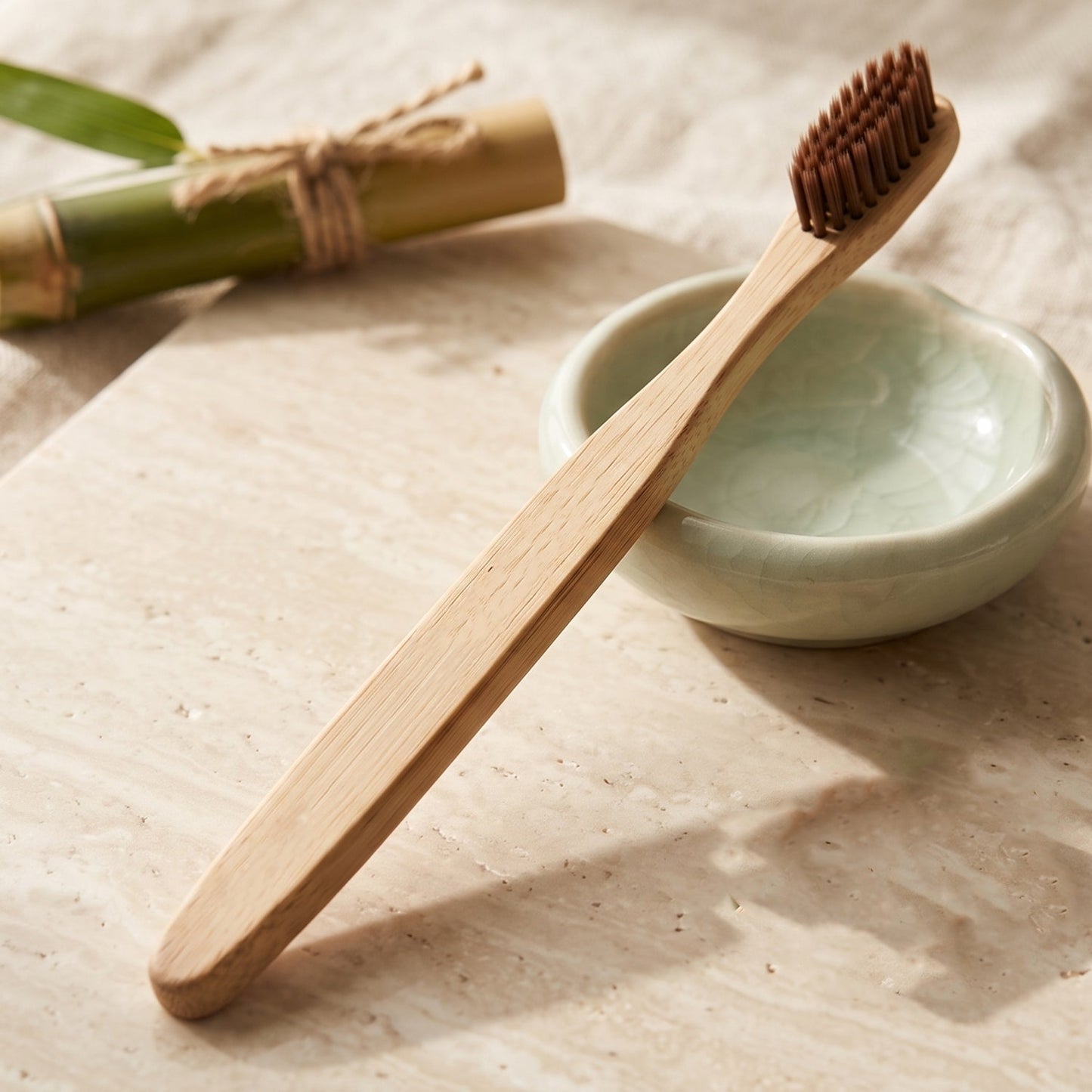 A bamboo toothbrush with brown bristles and an engraved handle rests on a light-colored surface next to a small green bowl