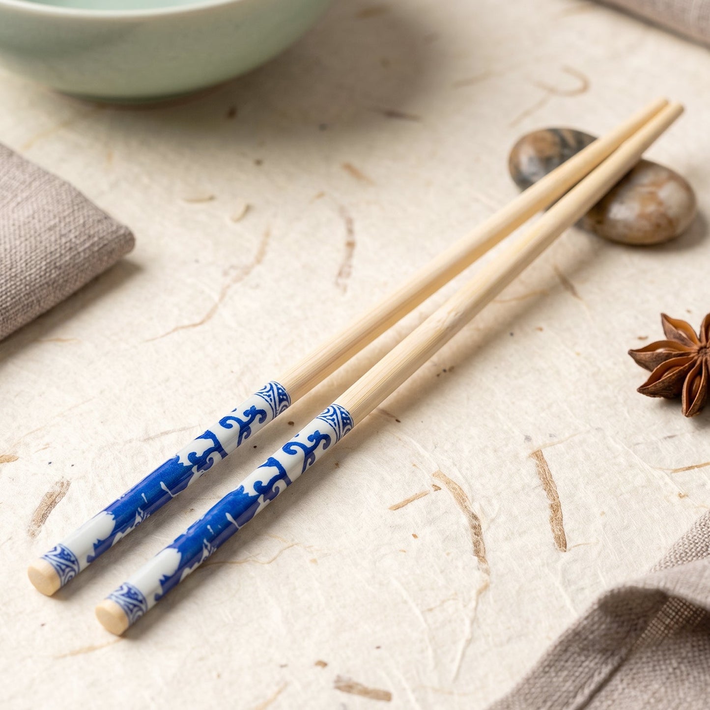 Two bamboo chopsticks with blue Chinese print rest on a textured white surface next to a light green bowl.