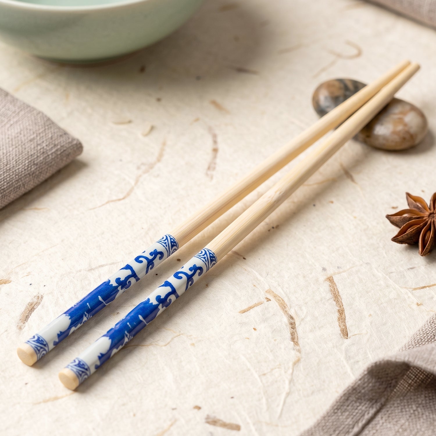 Two bamboo chopsticks with blue Chinese print rest on a textured white surface next to a light green bowl.