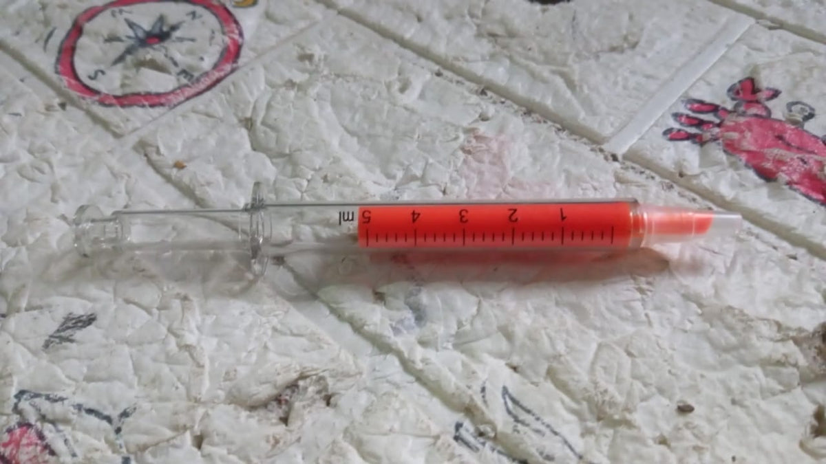 An orange syringe highlighter with a clear barrel and measurements, lying on a textured white and red surface.