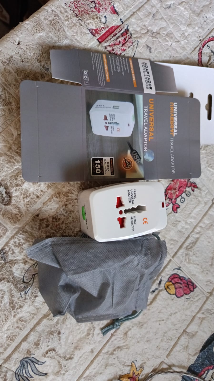 White universal travel adapter, its open box, and a gray drawstring pouch on a patterned bedspread.