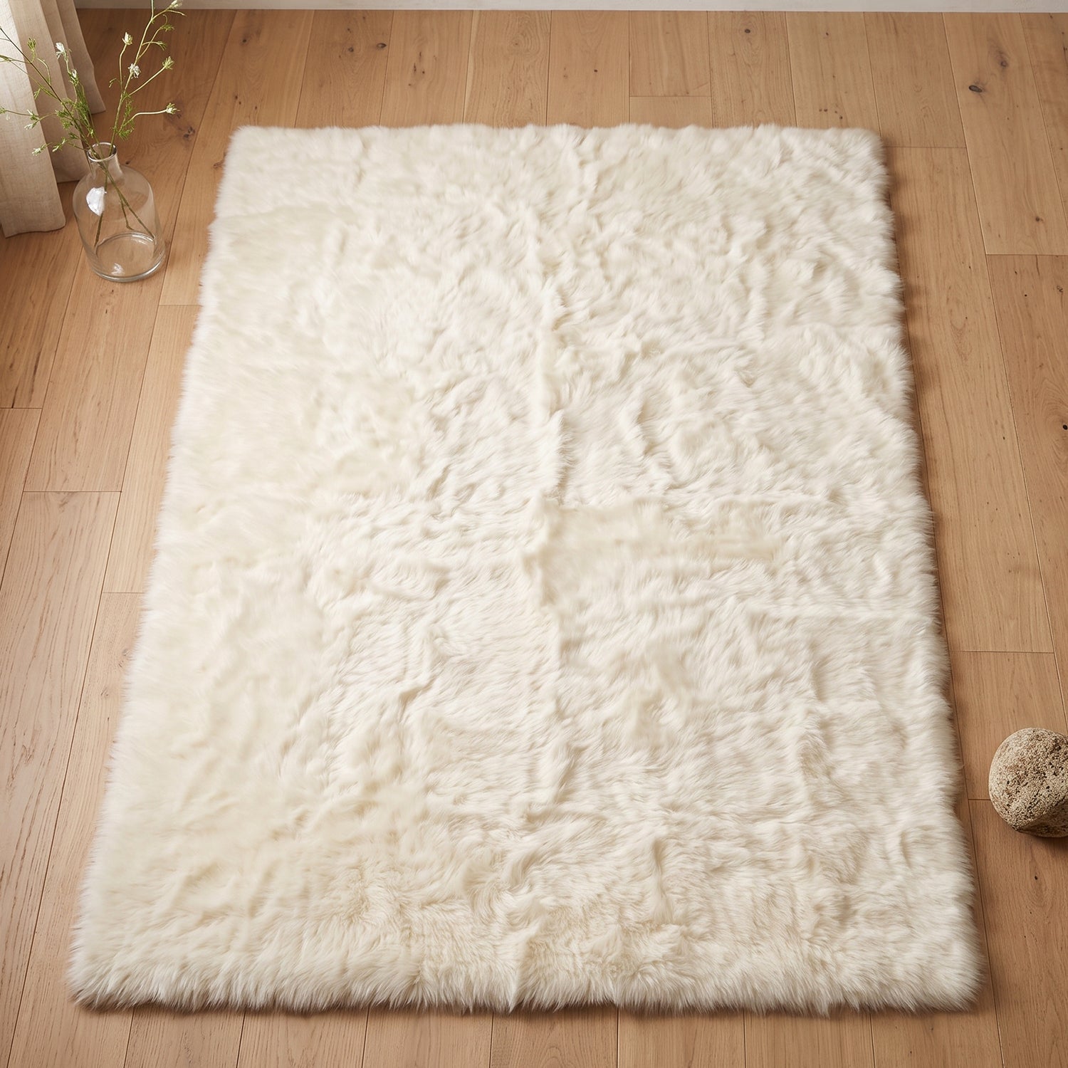A cream fluffy faux fur rug on a light wooden floor, with a small glass vase and a decorative rock.