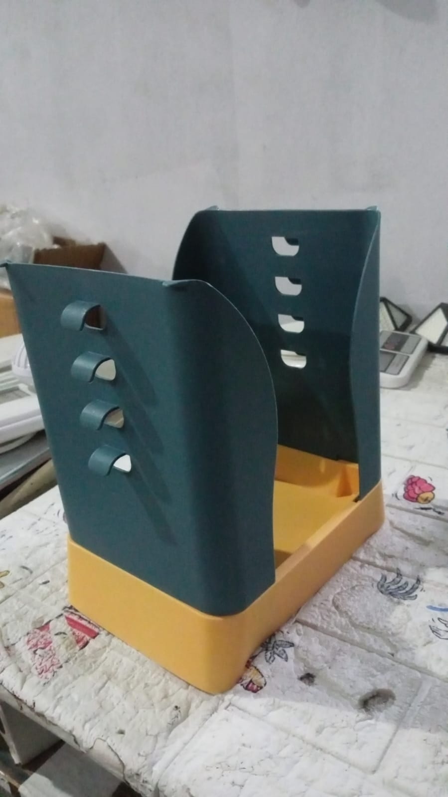 A teal and orange trash bag holder sits on a patterned surface in a room with various objects.