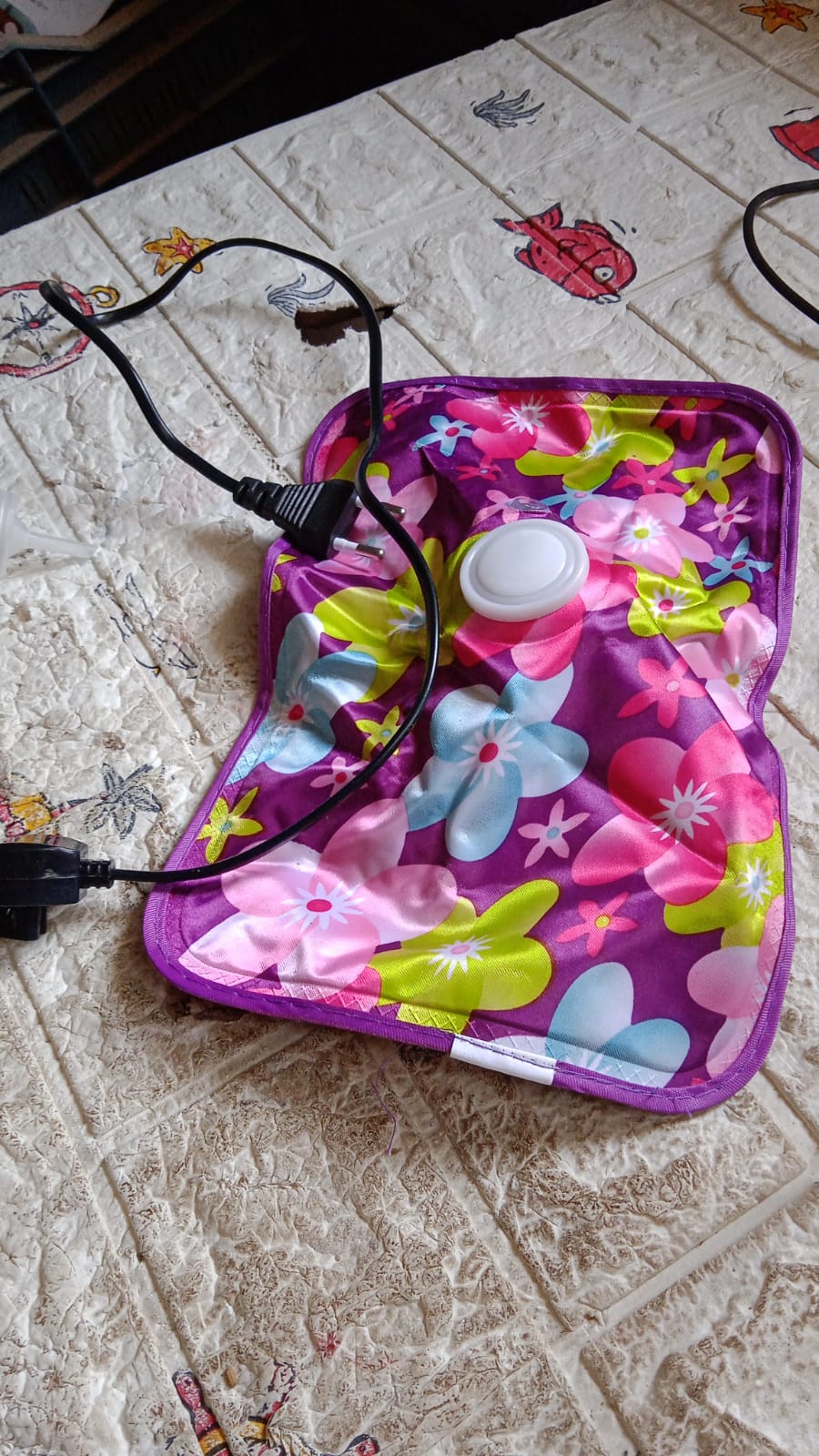 A purple floral hot water bag with colorful flowers is plugged in and lying on a textured light-colored surface.
