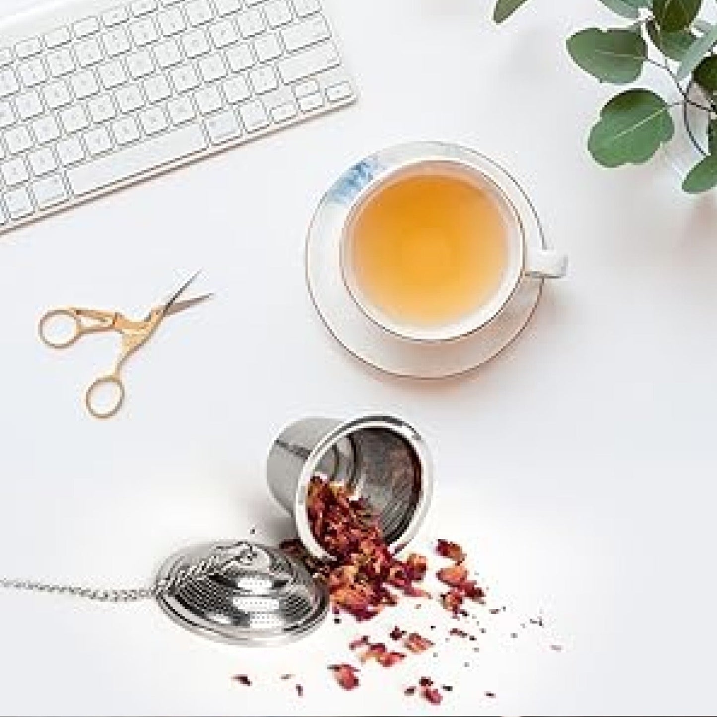 An overhead view of a stainless steel tea infuser with rose petals spilled out, a white teacup, and gold scissors on a white