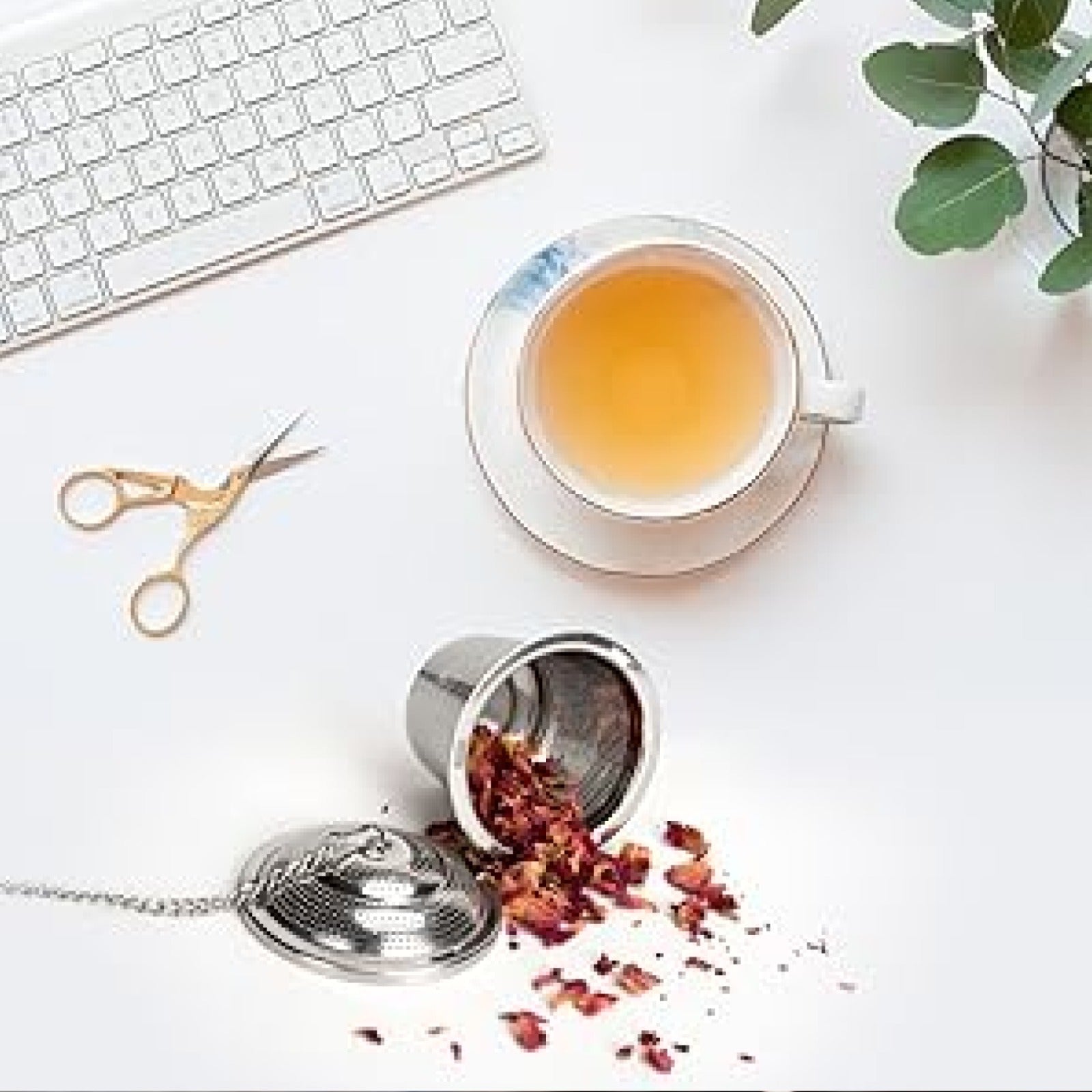 An overhead view of a stainless steel tea infuser with rose petals spilled out, a white teacup, and gold scissors on a white