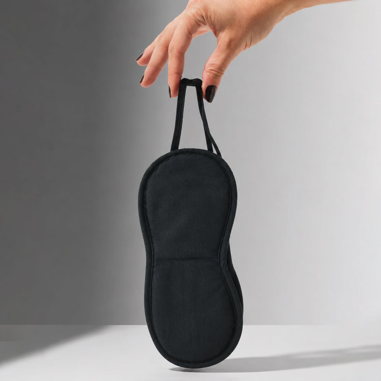A hand with dark nail polish holds up a black fabric sleep mask by its elastic strap against a grey background.