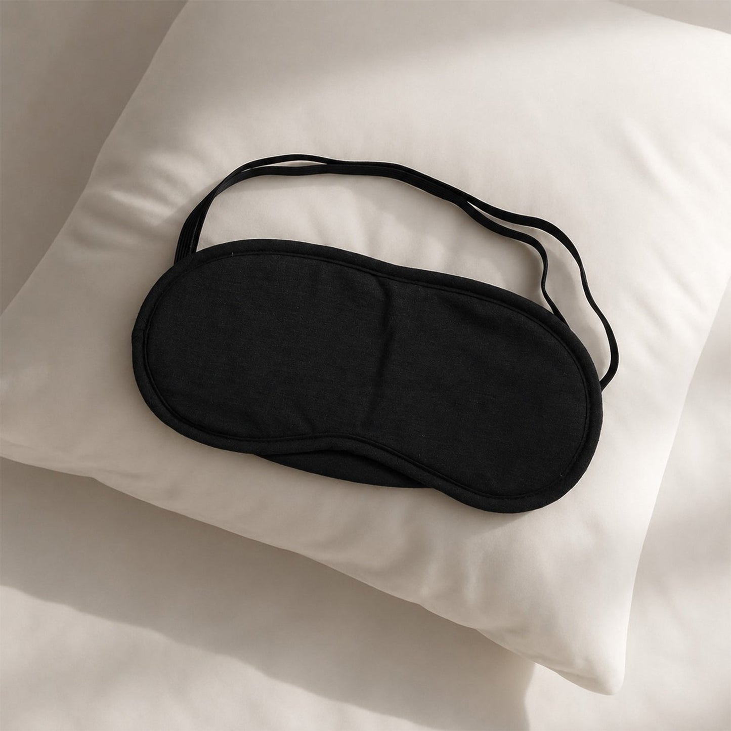 A soft black fabric sleeping mask with an elastic strap rests on a white pillow.