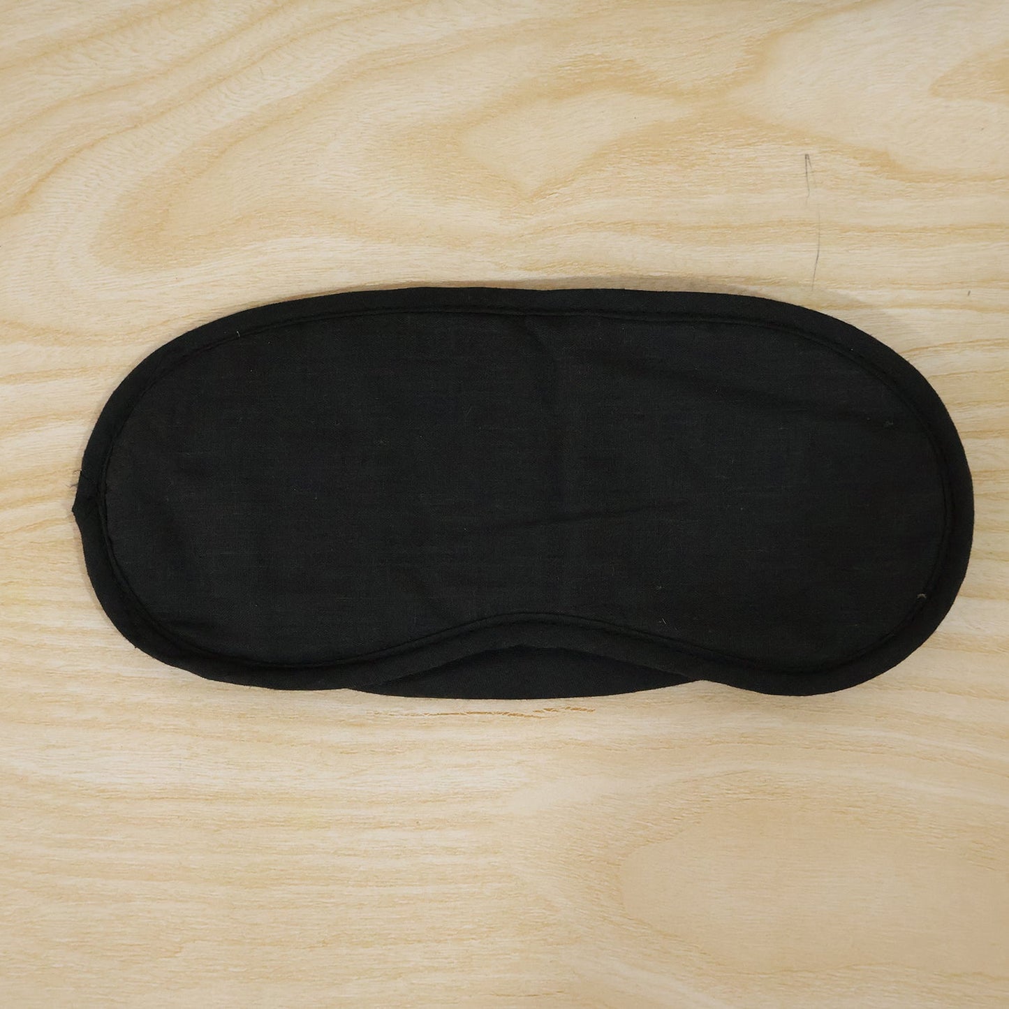 A black fabric sleeping mask with an elastic strap lies flat on a light wooden surface.