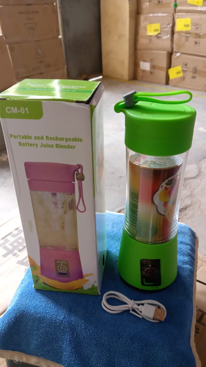 A green portable juicer blender with a white USB cable is next to its box on a blue towel, surrounded by cardboard boxes.