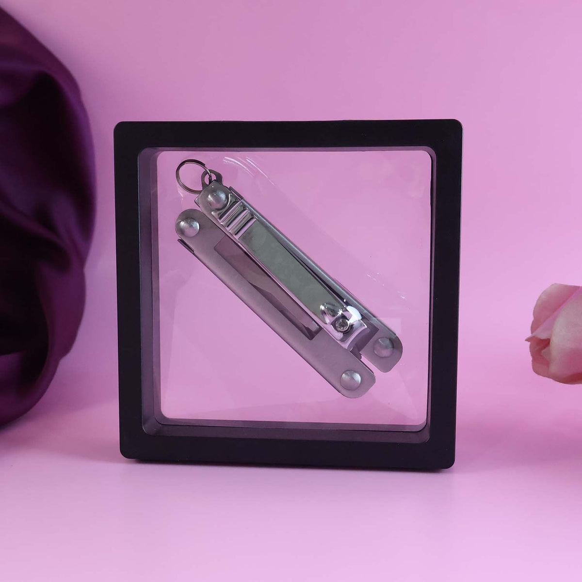 A folded silver multi-tool with a keyring in a clear display box, set against a pink background with a silk fabric.