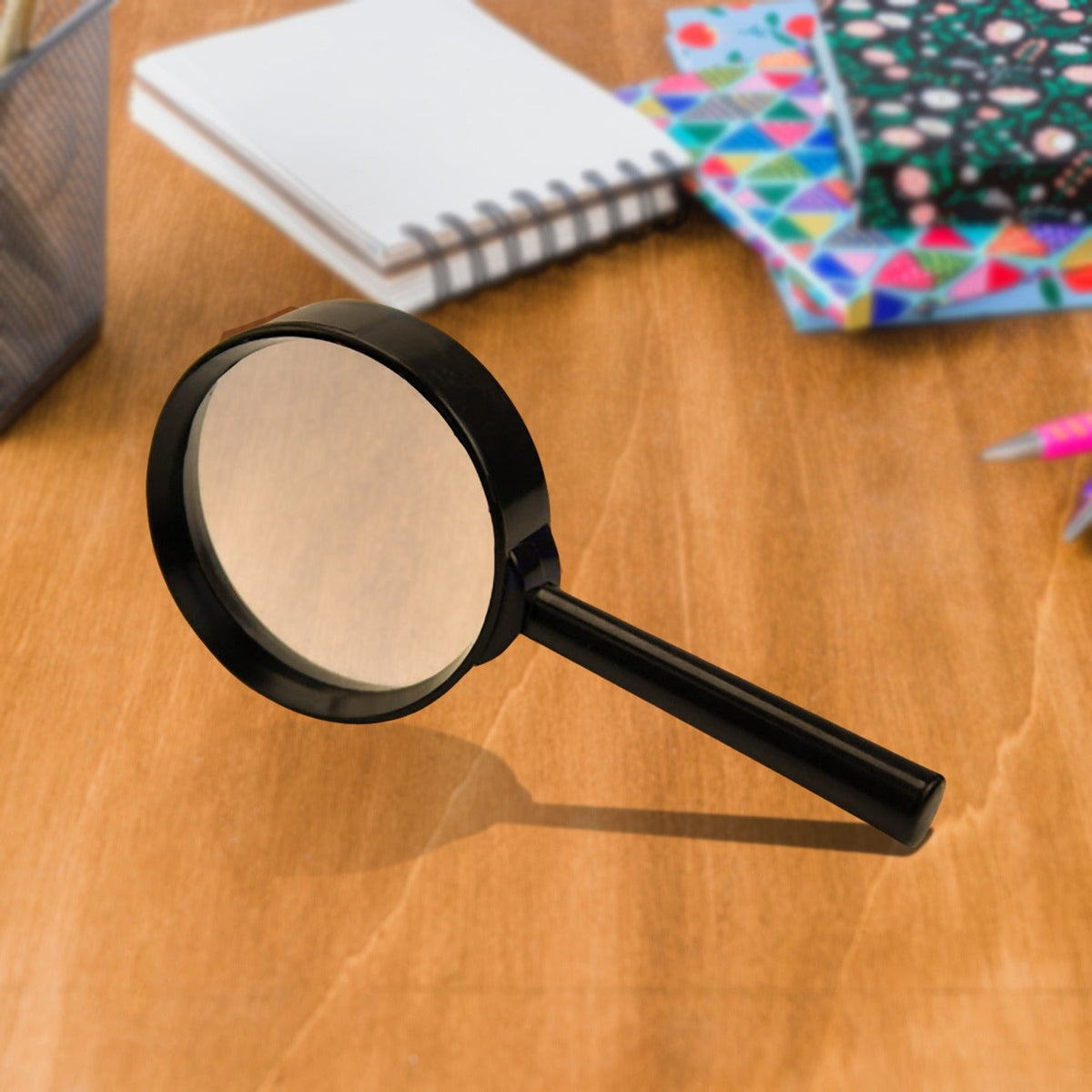 A black magnifying glass with a 50mm lens is positioned on a wooden desk, surrounded by office supplies.