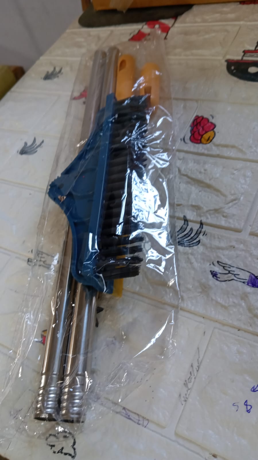 The blue and yellow floor cleaning brush, disassembled, is packaged in clear plastic on a patterned surface.
