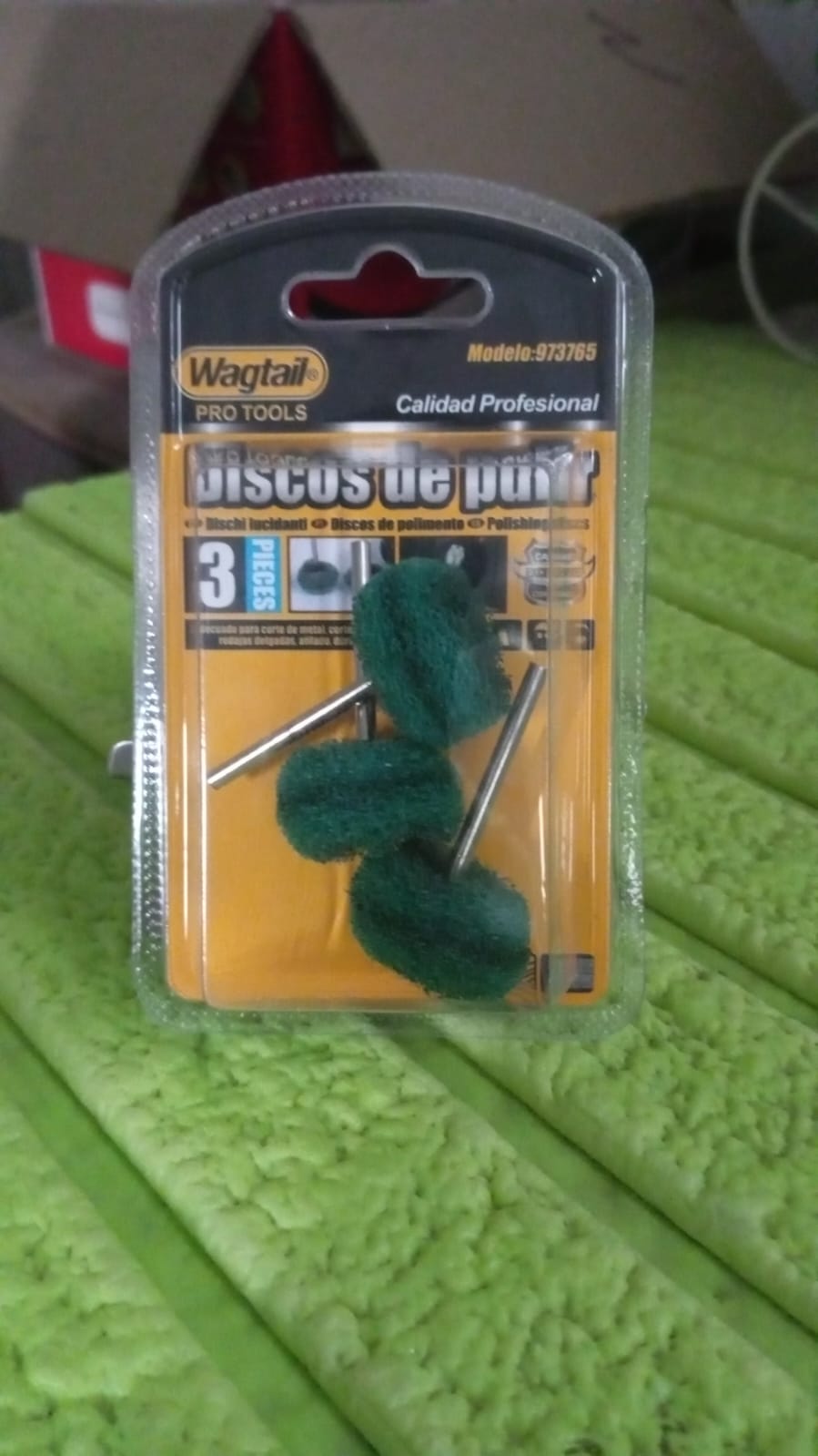 A blister pack containing three green polishing wheels with metal shanks, resting on a textured green surface.