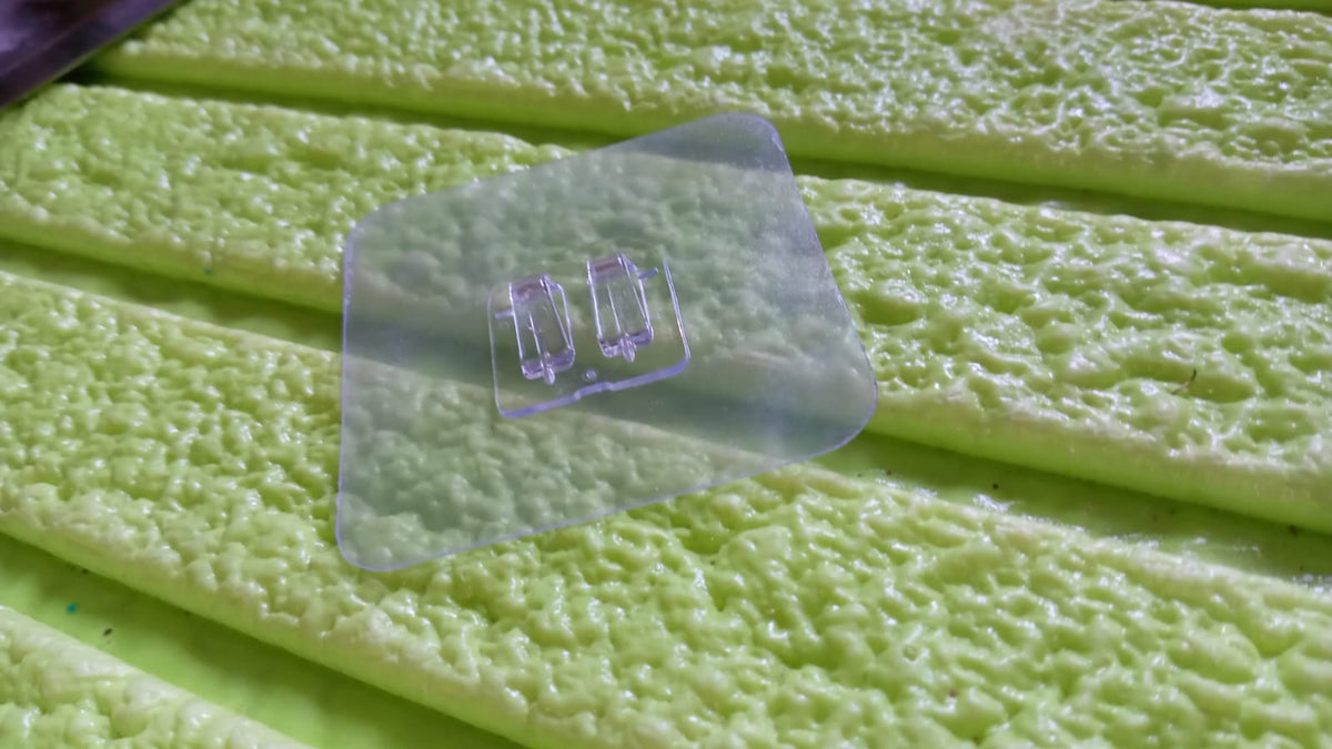 A clear self-adhesive wall hook with two prongs rests on a textured light green surface, possibly a cleaning pad.