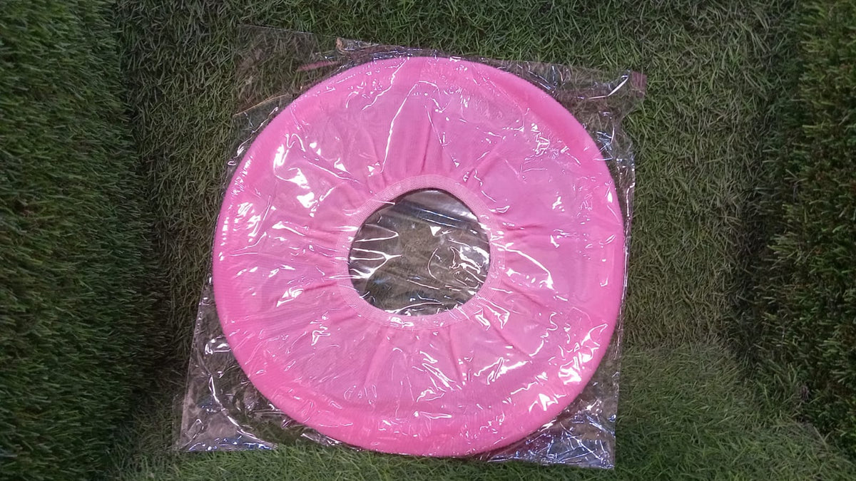A pink soft fabric toilet seat cover in clear plastic packaging resting on a green textured surface.