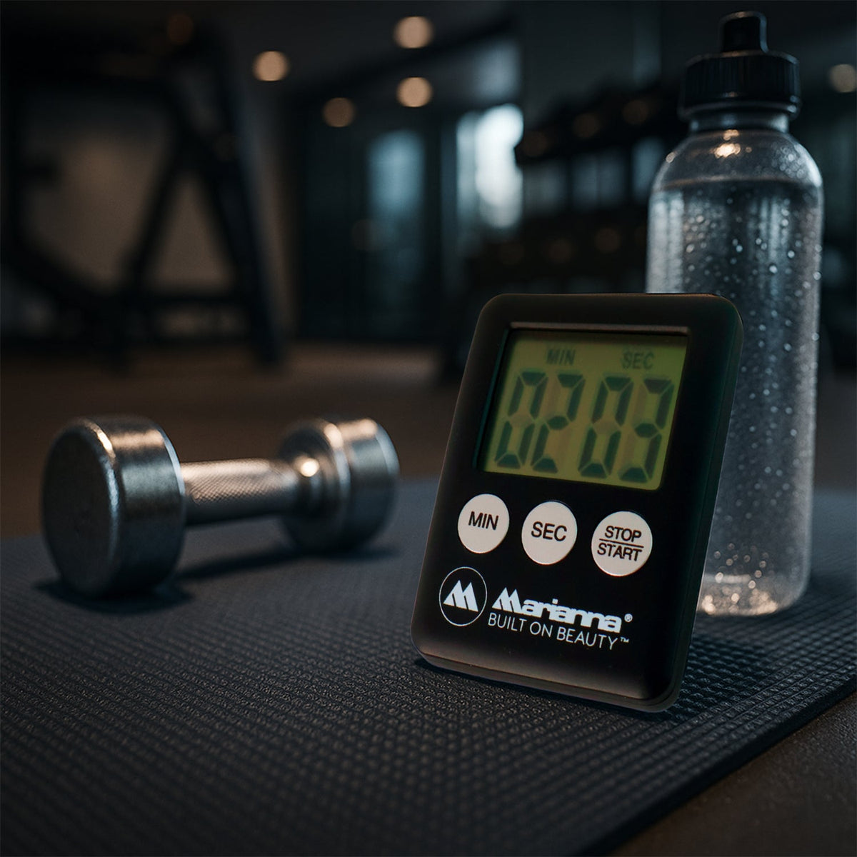 A black digital timer showing 02:03 sits on a black mat next to a dumbbell and a water bottle in a gym setting.