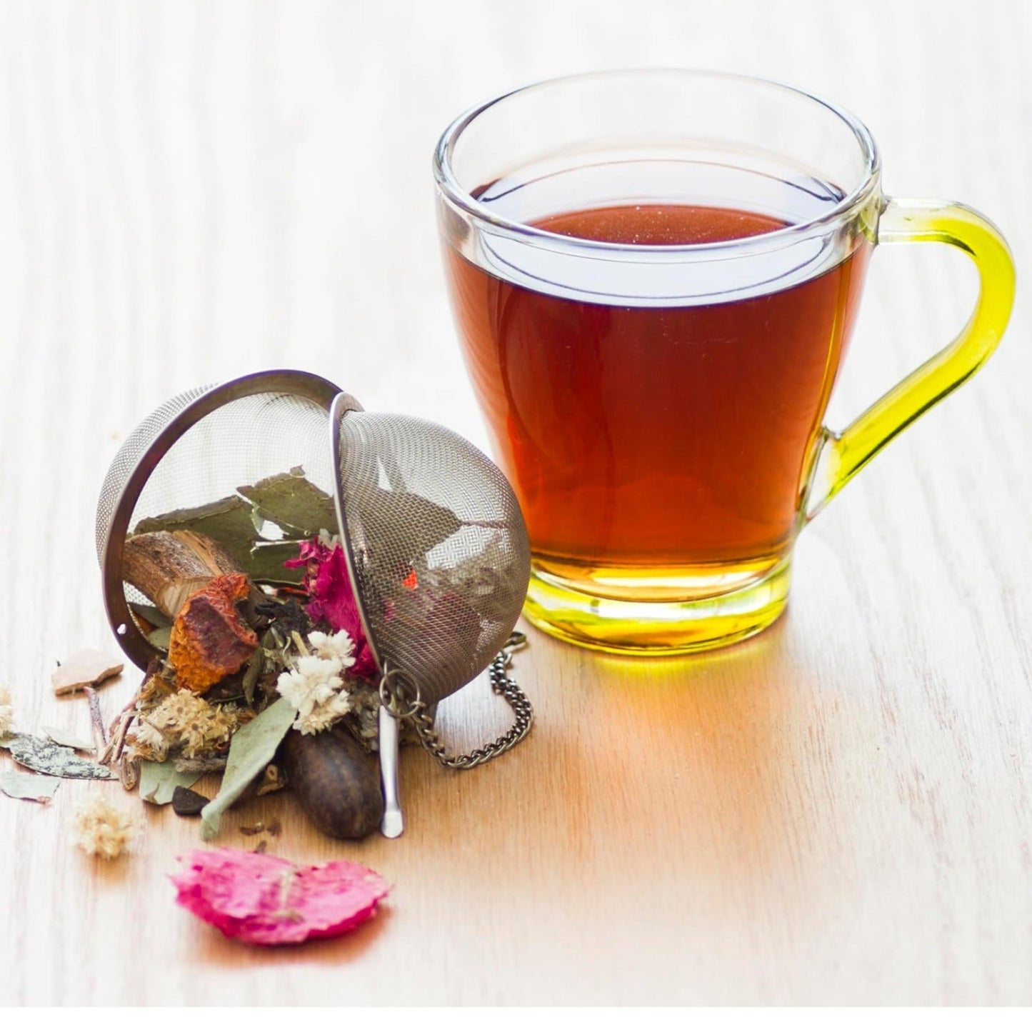 A stainless steel mesh tea infuser filled with dried flowers and herbs next to a clear glass mug of tea with a yellow handle.