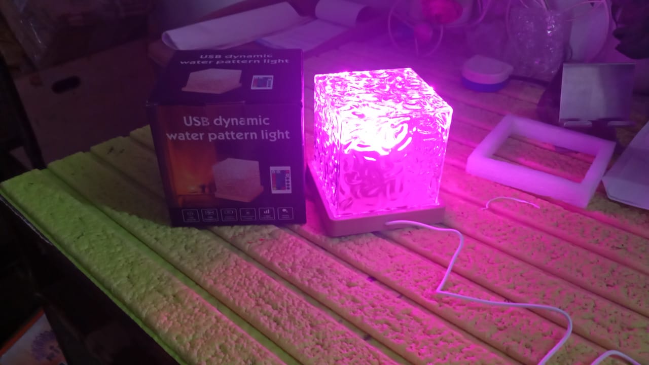 A crystal cube lamp projecting pink light, next to its product box, on a textured green surface.