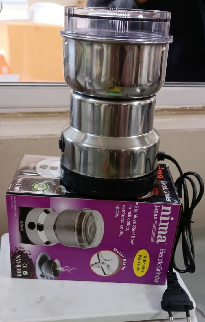 A stainless steel electric grinder sits on its purple and black product box, with the power cord visible.