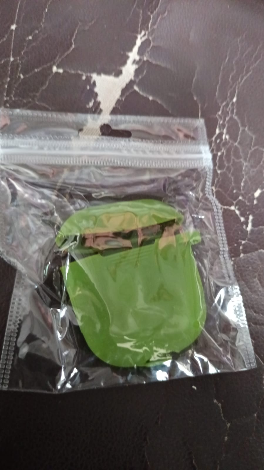 A green silicone earbud case is sealed inside a clear plastic bag on a dark brown textured surface.