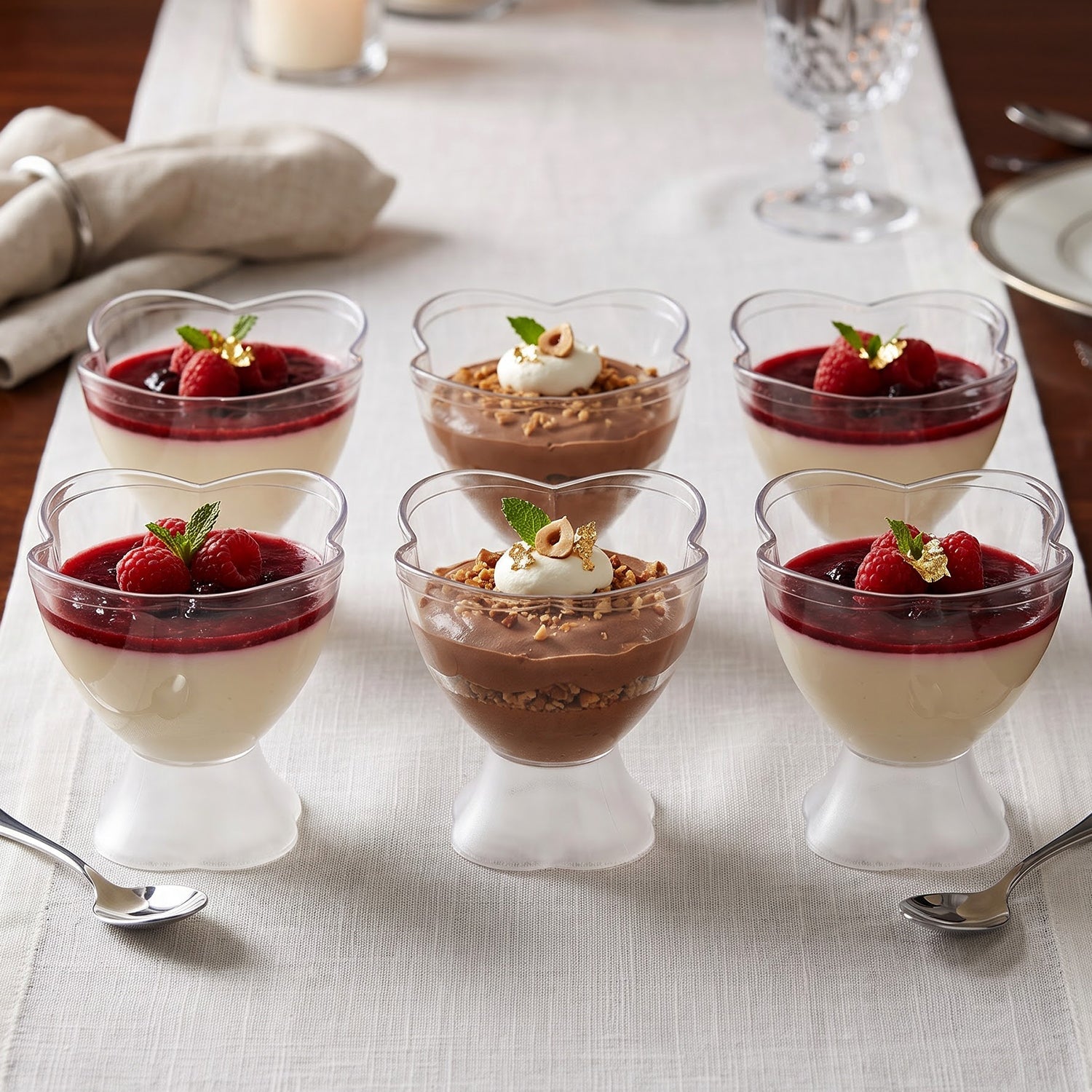 Six clear plastic dessert bowls with heart-shaped rims, filled with various desserts, on a white table runner, with spoons.