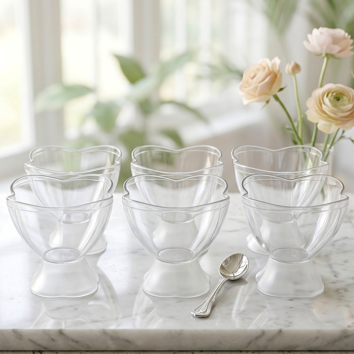 Six empty clear plastic dessert bowls with heart-shaped rims and frosted bases on a marble counter with a spoon and flowers.