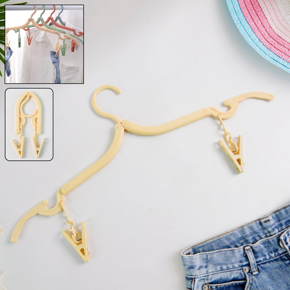 A beige foldable clothes hanger with two clips lies on a white surface next to blue jeans and a hat