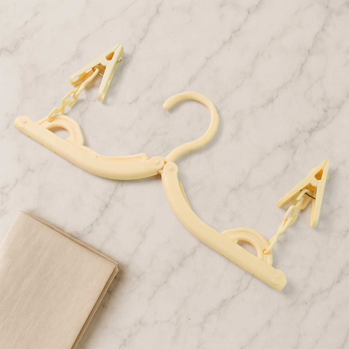 A beige foldable clothes hanger with two clips lies partially folded on a white marble surface next to a folded beige cloth.