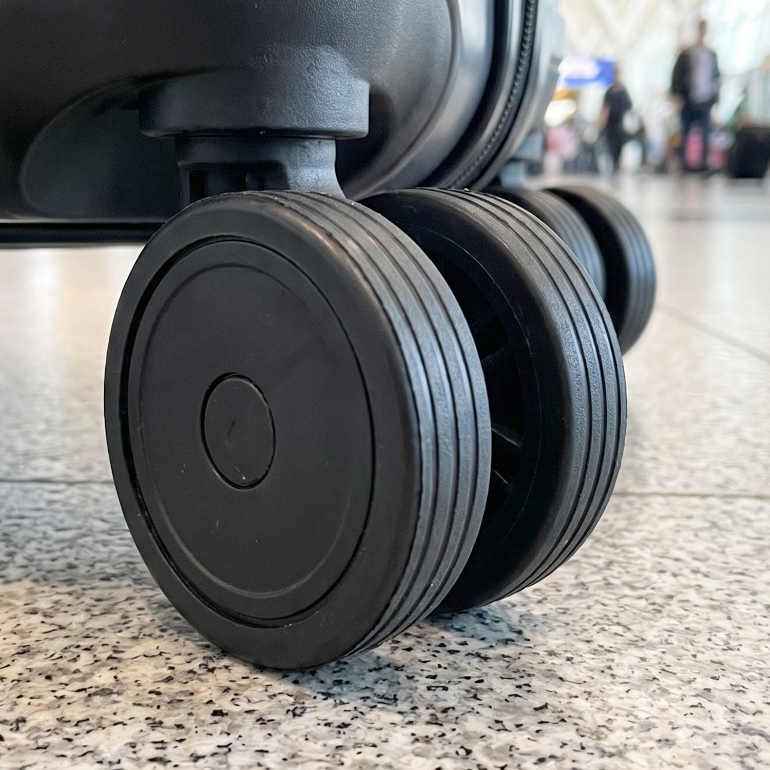 Three black luggage wheels on a grey tiled floor, with a blurred background of an airport terminal.
