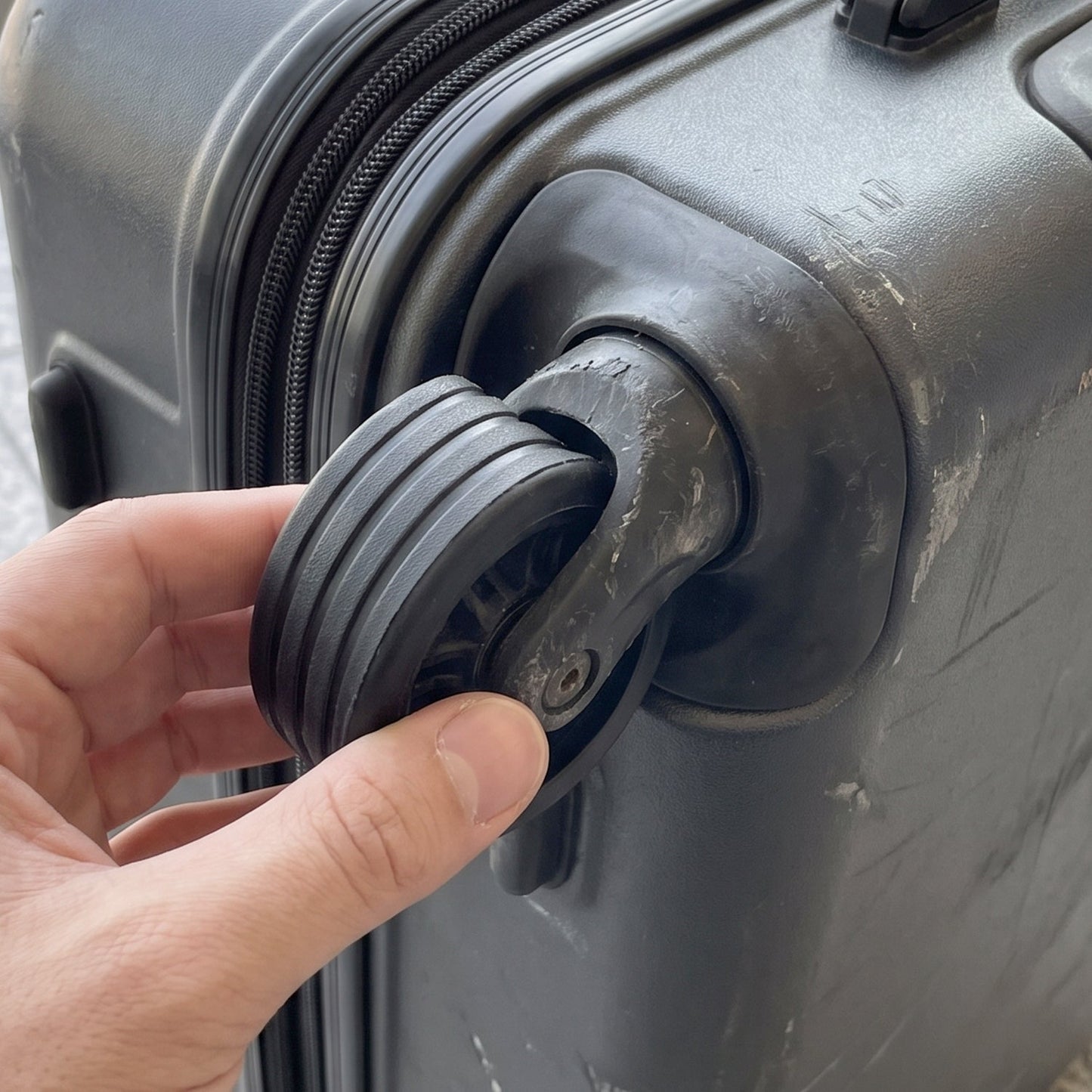 A hand holding a black silicone ring around a luggage wheel, demonstrating its application on a grey suitcase.