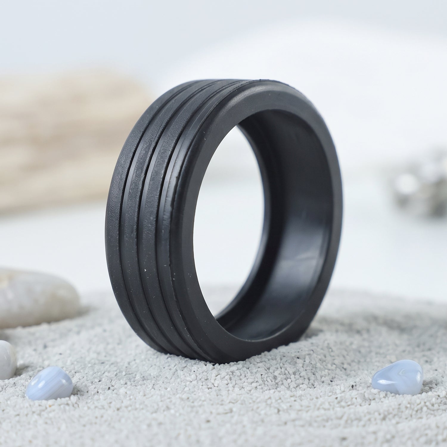 A single black silicone ring with three raised ridges, standing upright on light sand with small white and blue pebbles.