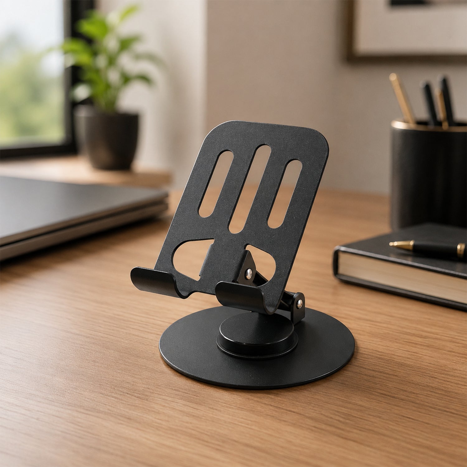 A black metal mobile stand, with a slotted back, sits on a wooden desk next to a notebook and pen holder.