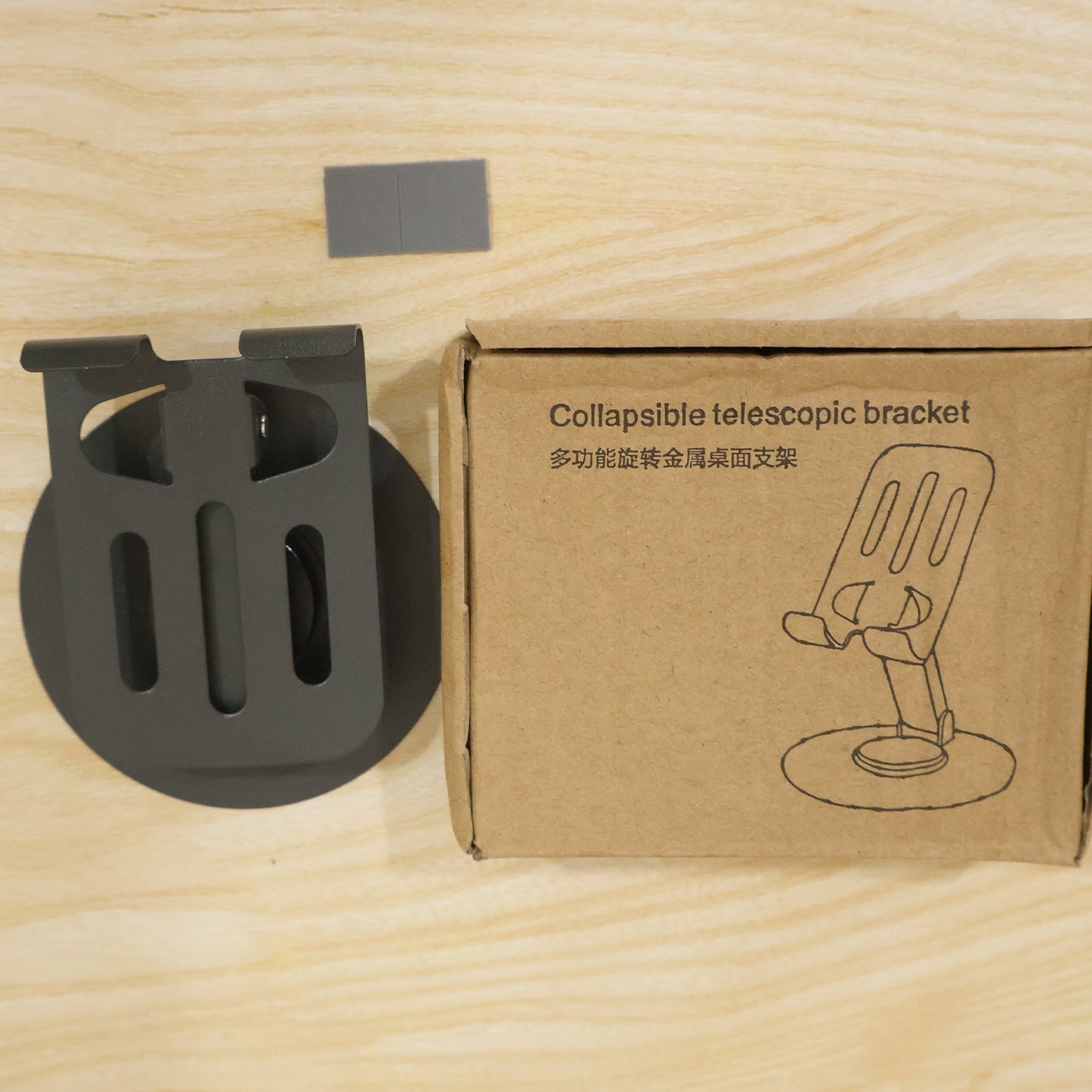 A black metal mobile stand next to its brown cardboard box labeled "Collapsible telescopic bracket" on a wooden surface.