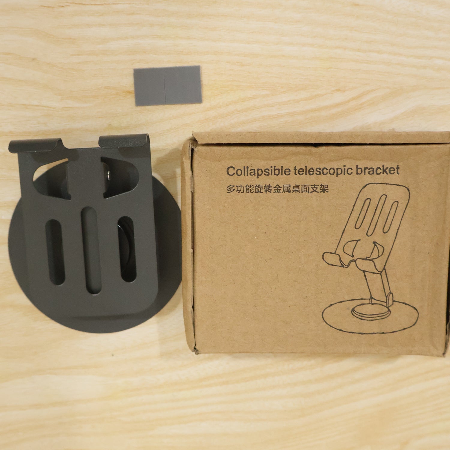 A black metal mobile stand next to its brown cardboard box labeled "Collapsible telescopic bracket" on a wooden surface.