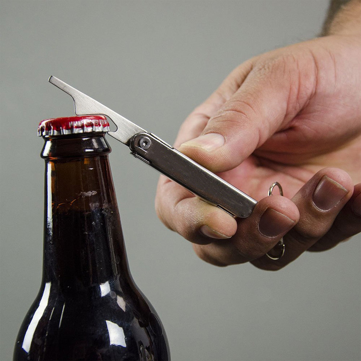 A hand uses the silver multi-tool to open a brown glass bottle with a red cap.