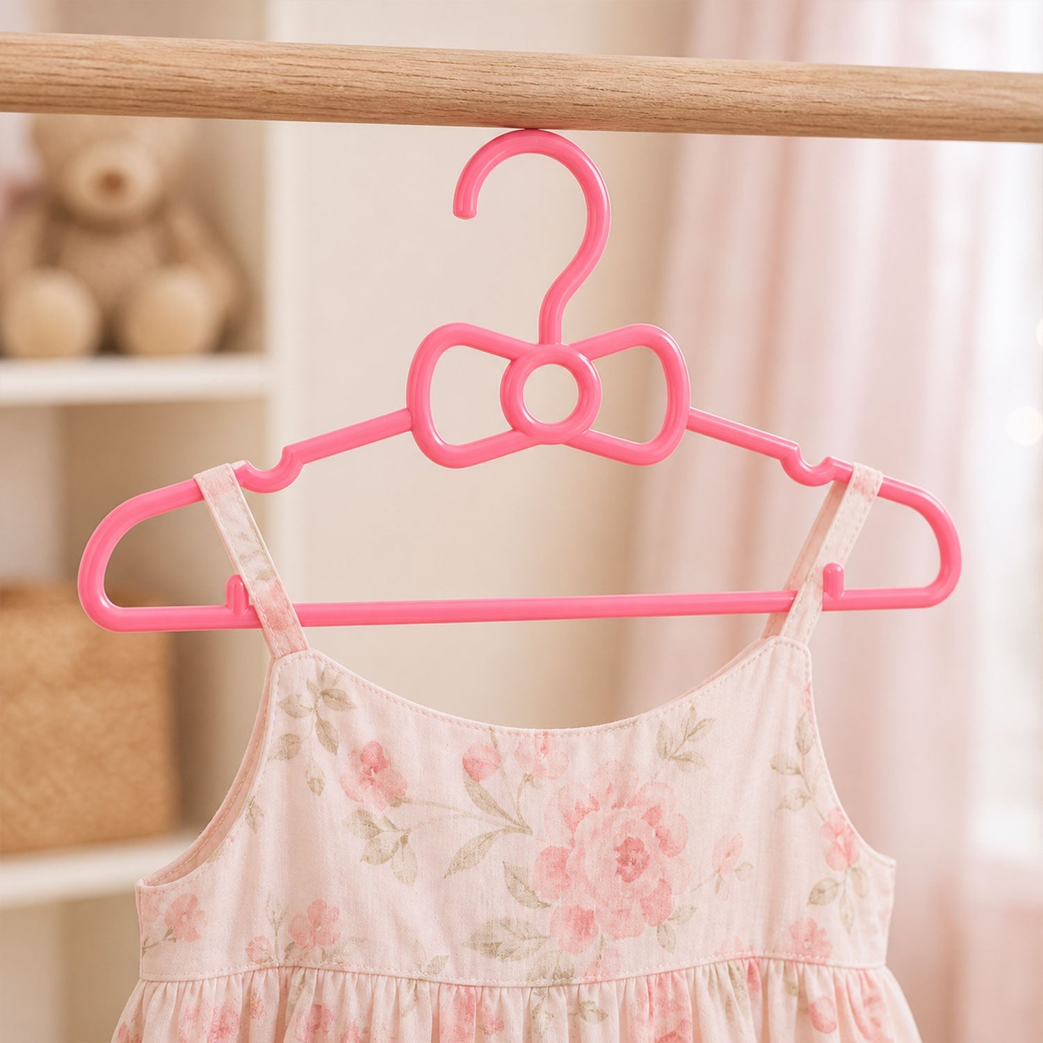 A pink floral baby dress hangs on a pink plastic bow-shaped hanger from a wooden rod.