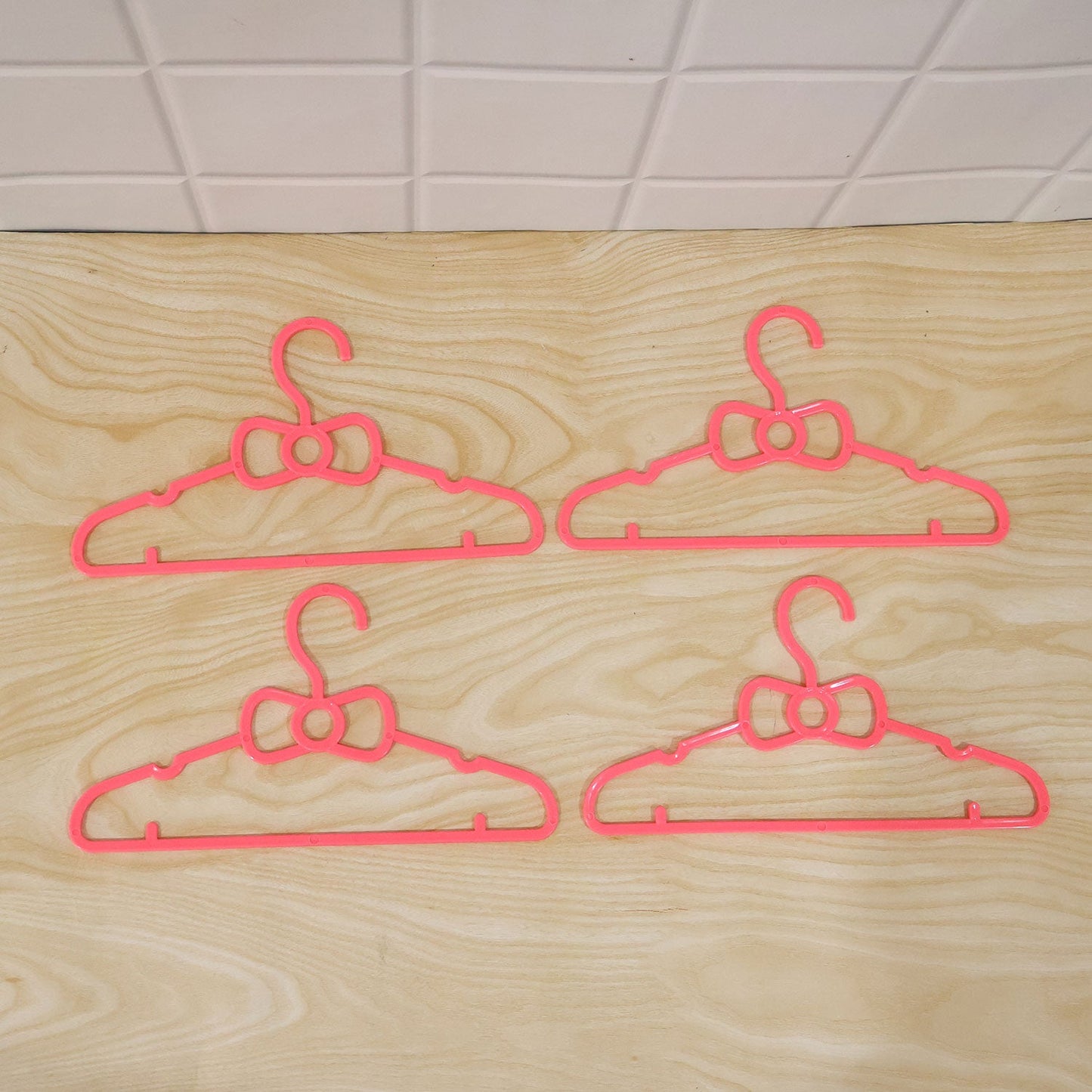 Four pink plastic bow-shaped hangers are arranged on a light wooden surface with a tiled background.