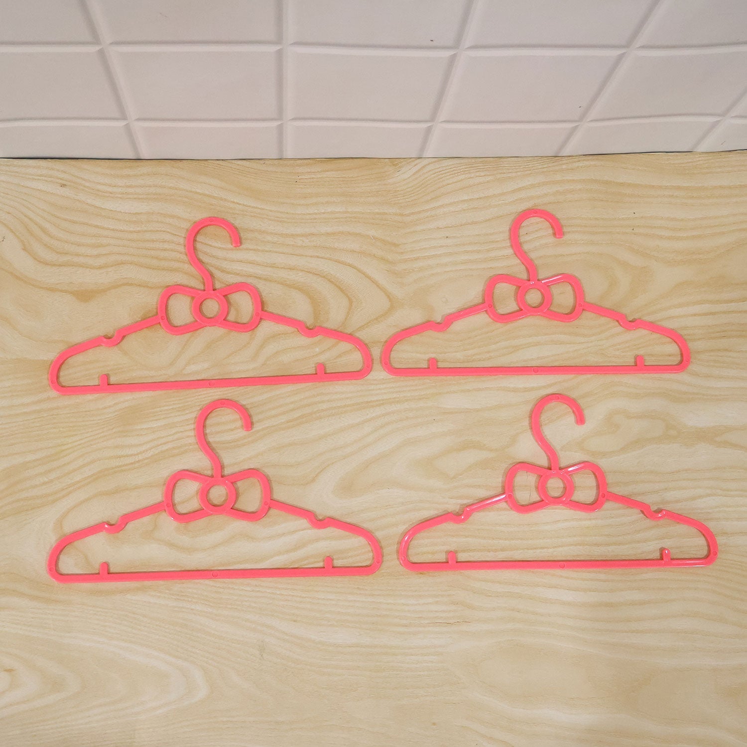 Four pink plastic bow-shaped hangers are arranged on a light wooden surface with a tiled background.