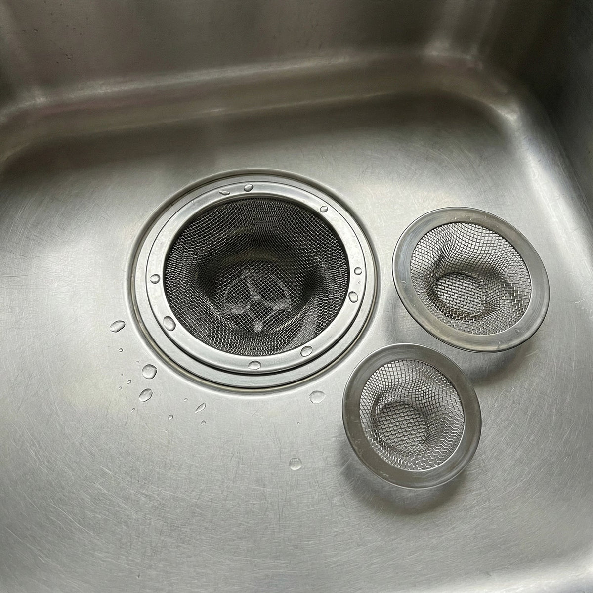 Three stainless steel mesh sink strainers in a stainless steel sink with water droplets.