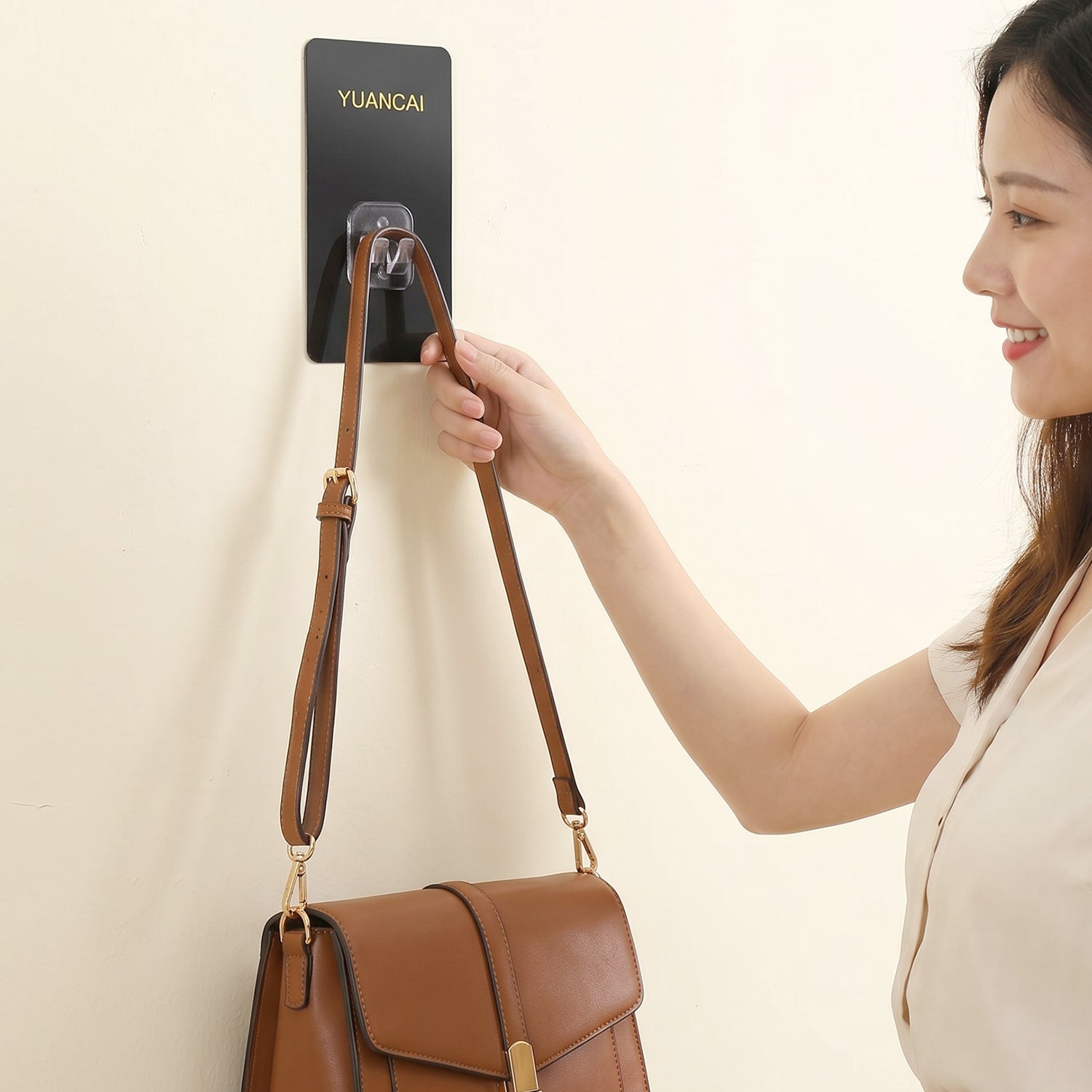 A woman's hand reaches to adjust a brown leather shoulder bag hanging from a black adhesive wall hook.