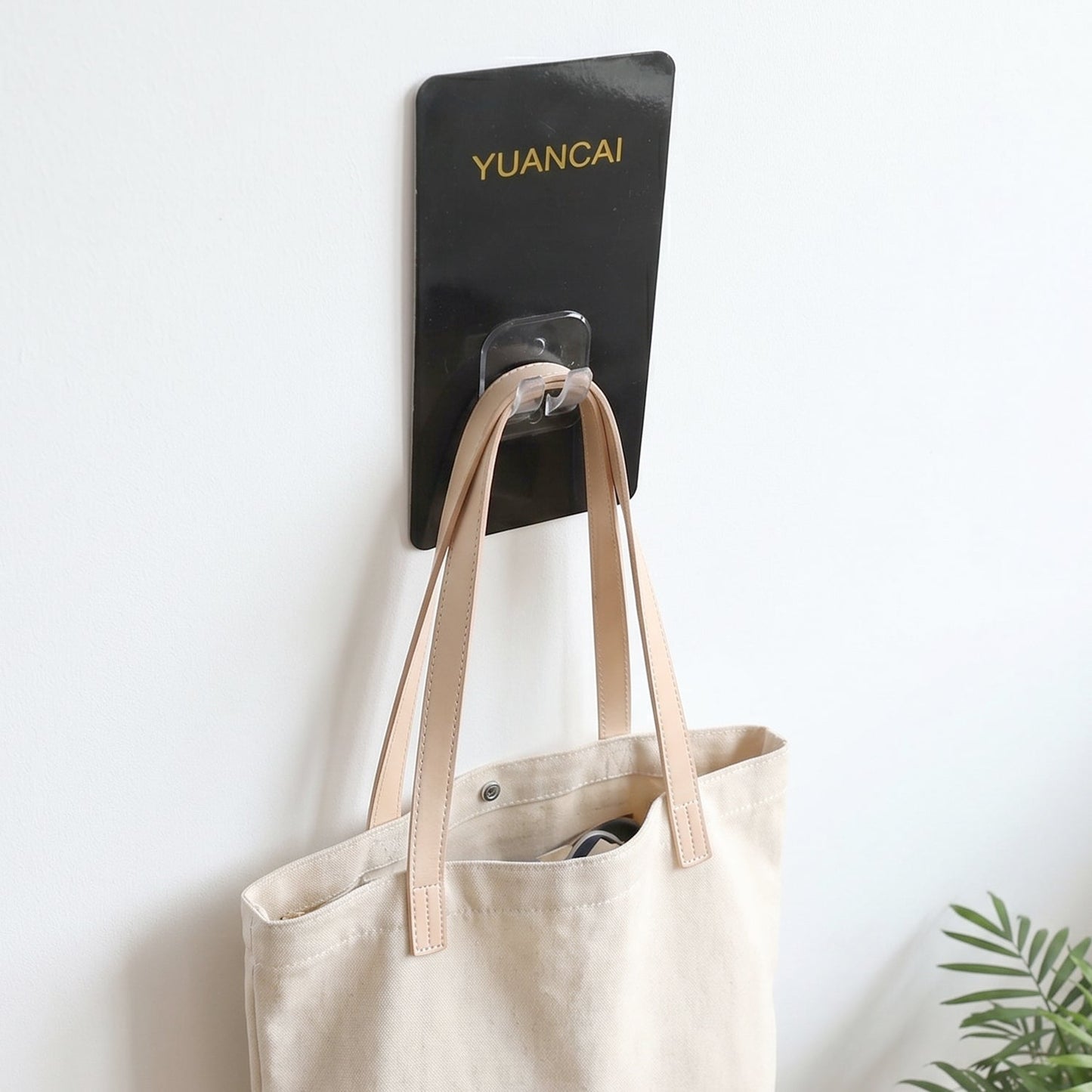 A cream-colored canvas tote bag with light brown leather straps hangs from a black adhesive wall hook.